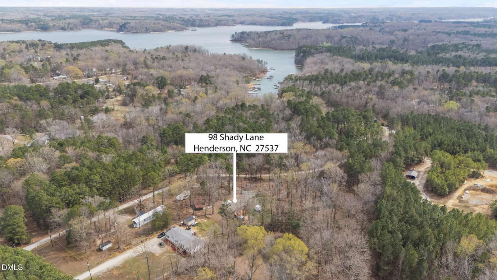 Aerial view of a wooded area near a lake with houses scattered among trees. A label with an arrow points to a property marked "98 Shady Lane, Henderson, NC 27537." The lake and forested landscape are visible in the background.