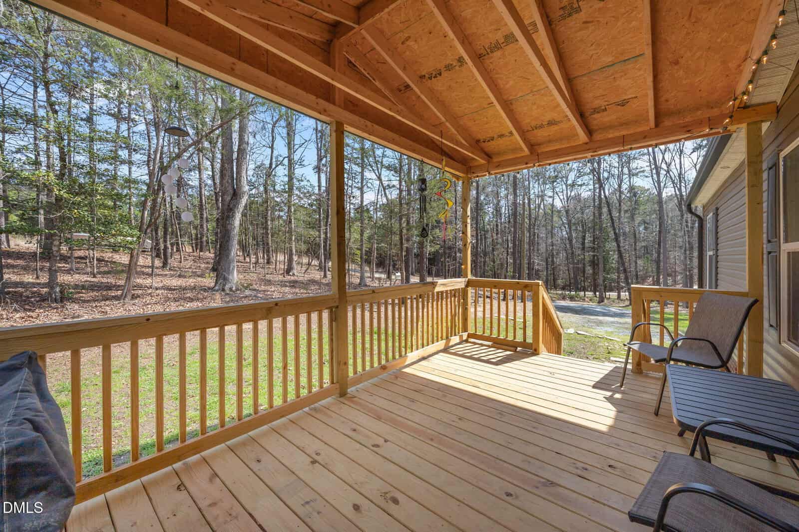 Covered wooden porch with two chairs overlooks a backyard with grass and trees. Sunlight filters through the roof, and the peaceful forested area is visible beyond the railing.