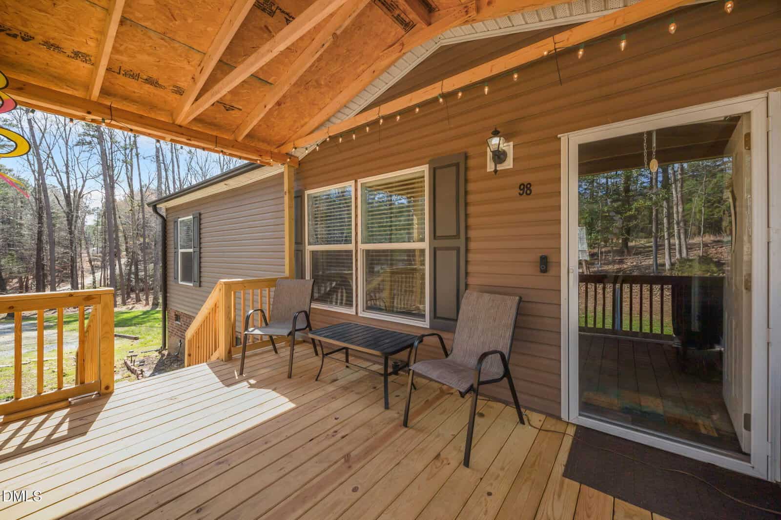 Covered wooden porch with two chairs, a table, and string lights. Brown siding house with large windows and a sliding glass door. Trees and grass visible in the background.