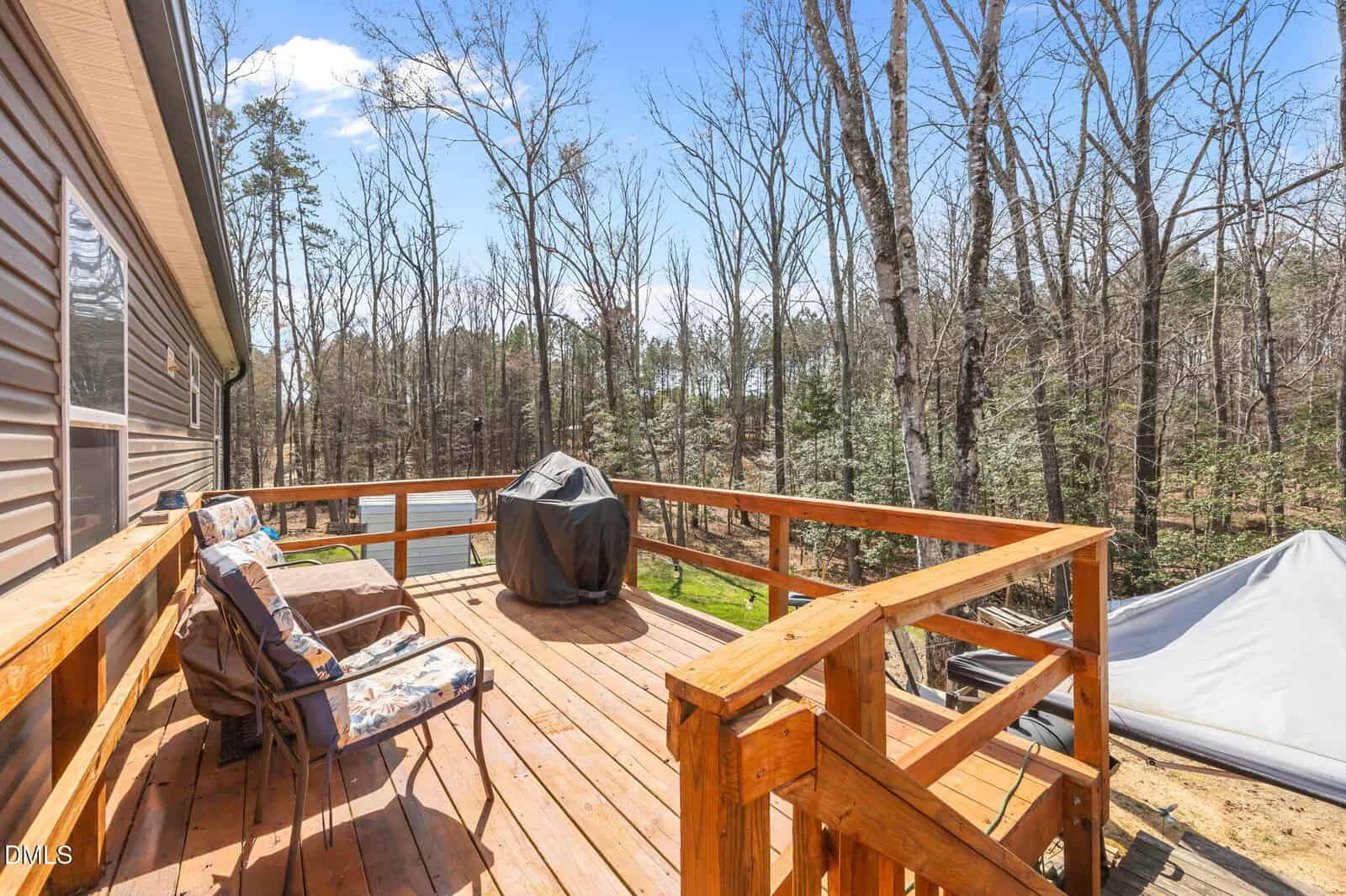 Wooden deck with railings, two cushioned chairs, and a grill covered with a black cover. Surrounded by leafless trees under a clear blue sky. A tent is visible off to the side.