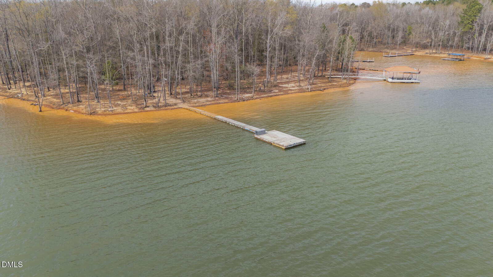 A long wooden dock extends from a sandy, tree-lined shoreline into a calm lake, with a covered boat dock visible in the background among bare trees.