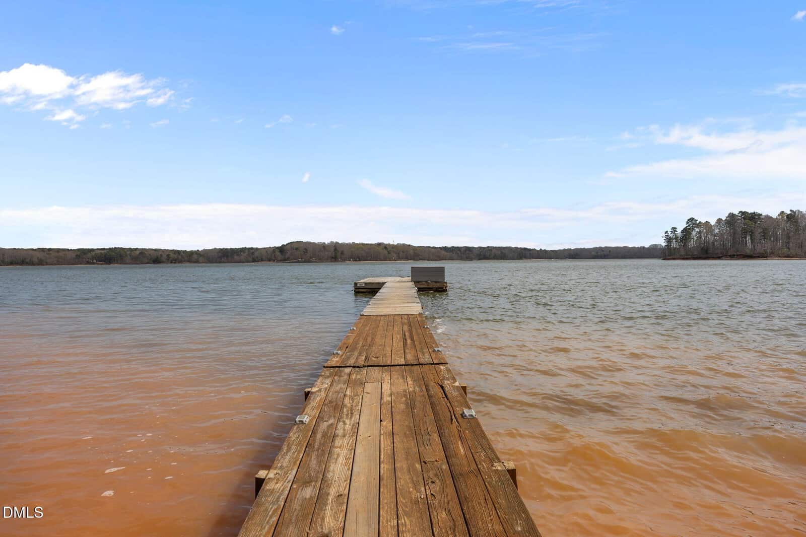 A wooden dock extends out over a large lake at 98 Shady Lane in Henderson, with calm water and a distant tree-lined shoreline under a blue sky with scattered clouds.