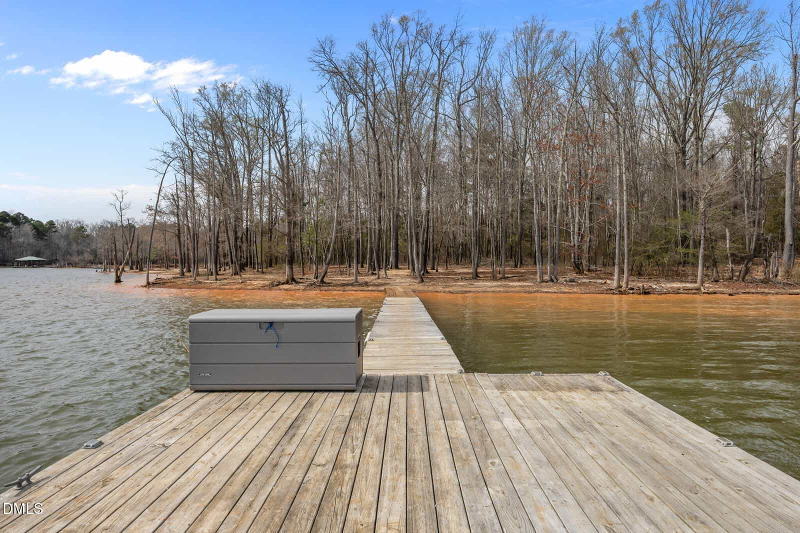 A wooden dock stretches over a calm lake toward a shoreline with leafless trees. A gray storage box sits on the dock under a blue sky with scattered clouds.