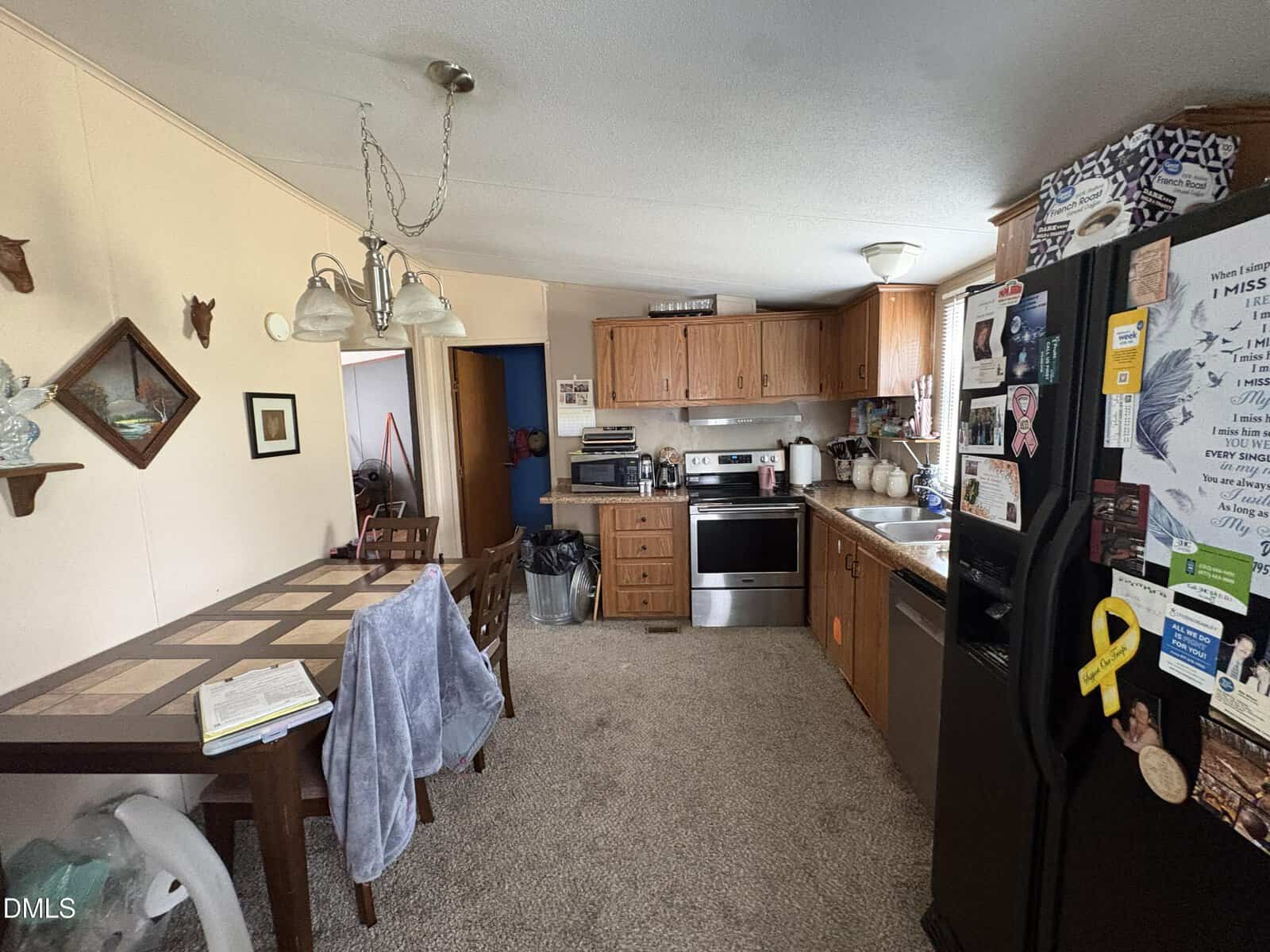 A cozy kitchen and dining area with wood cabinets, a black refrigerator covered in magnets, a stove, and a dishwasher. A dining table with chairs and a gray towel is in the foreground, with a notebook and mug on it.