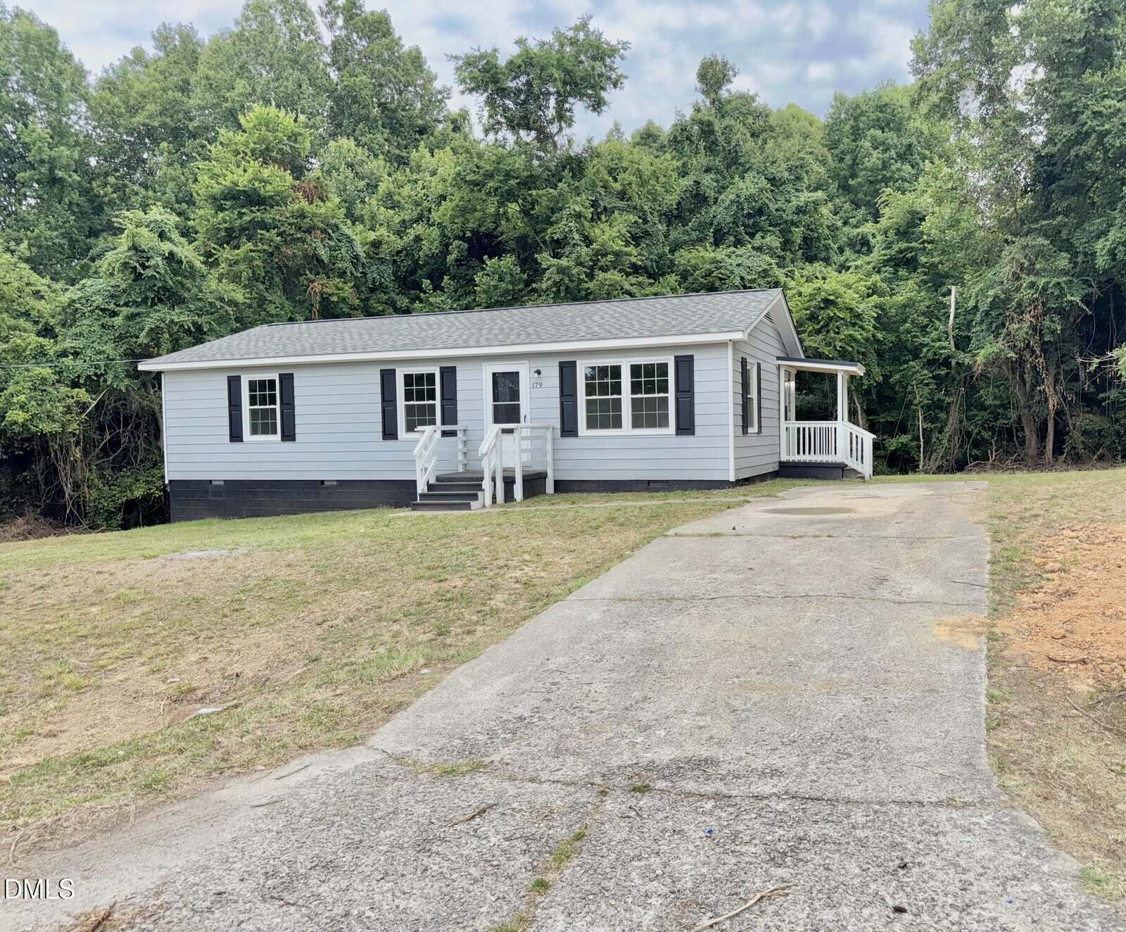 Single-story light gray house with black shutters, front porch, and white railings. The house sits on a grassy lot with a long concrete driveway and is surrounded by trees in the background.