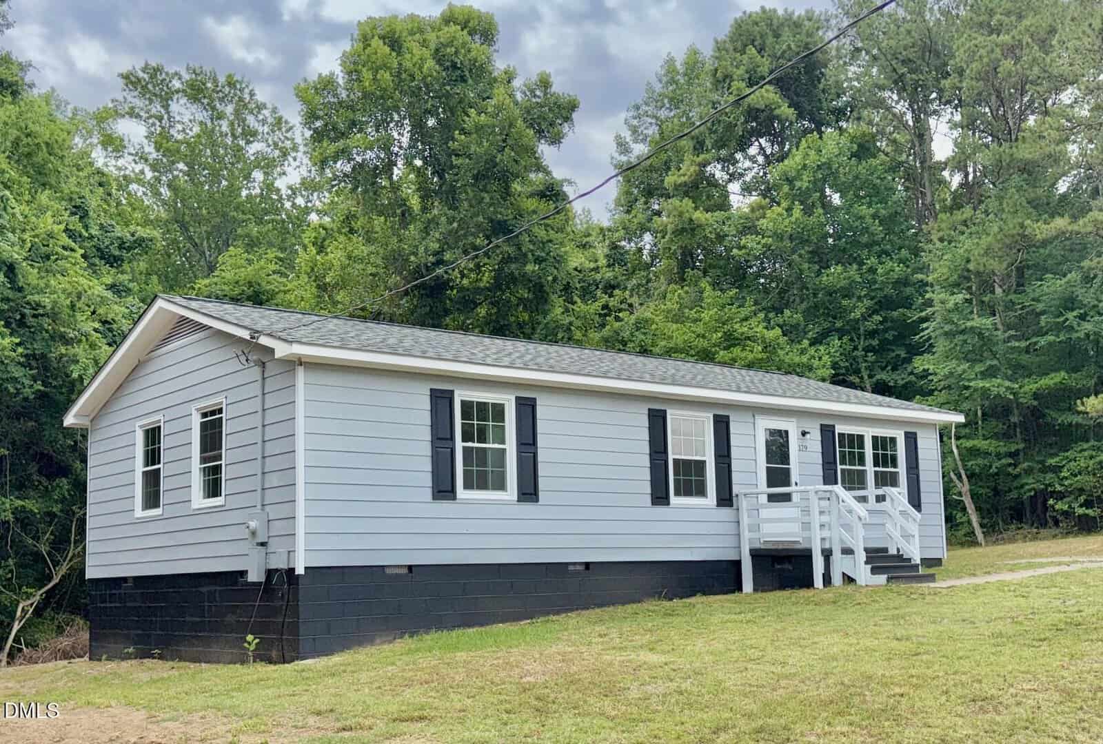 A light gray single-story house with black shutters and a small front porch sits on a grassy lot at 179 Lynnbank Estates Road in Kittrell, surrounded by tall green trees under a partly cloudy sky.