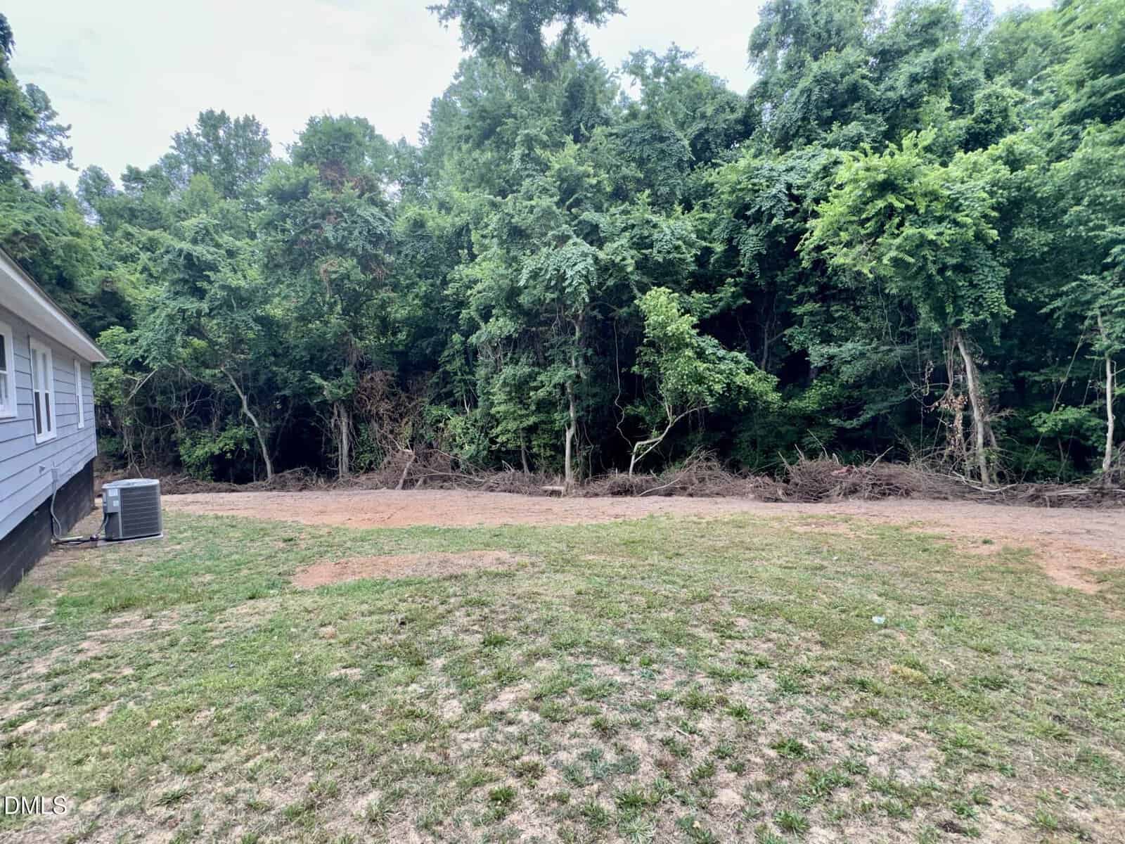 A patchy grass yard with a gray house on the left and dense green trees forming a wooded area in the background. An outdoor AC unit is visible beside the house.