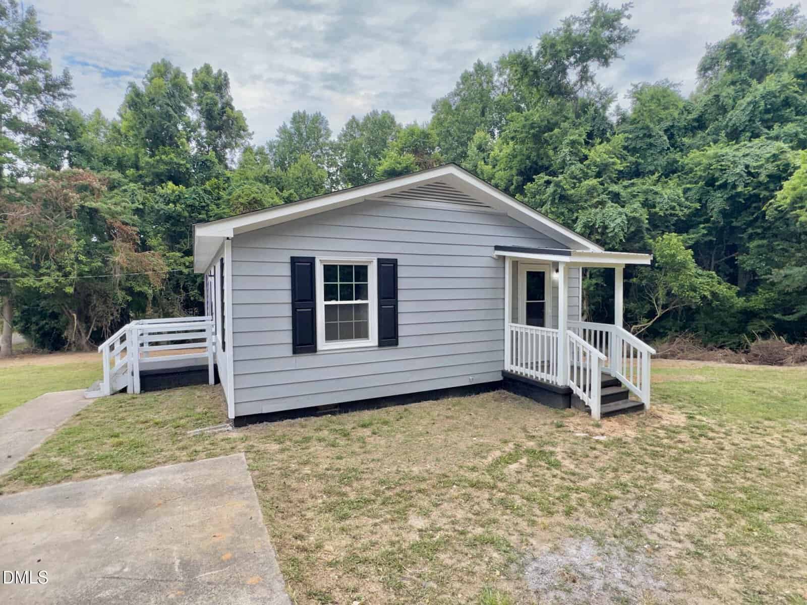A small gray house with white trim and black shutters, featuring a covered front porch with railings, sits on a grassy lot with surrounding trees and a wooden deck on one side.