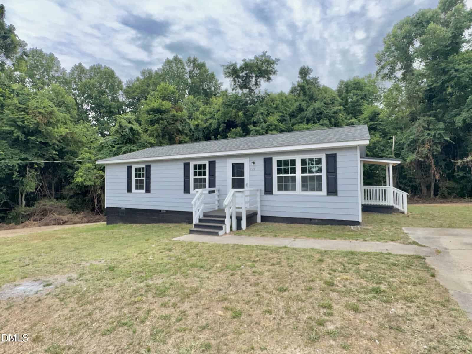 A single-story light blue house with white trim and black shutters, set on a grassy lot with trees in the background. The house has a front porch with white railings and steps leading to the front door.