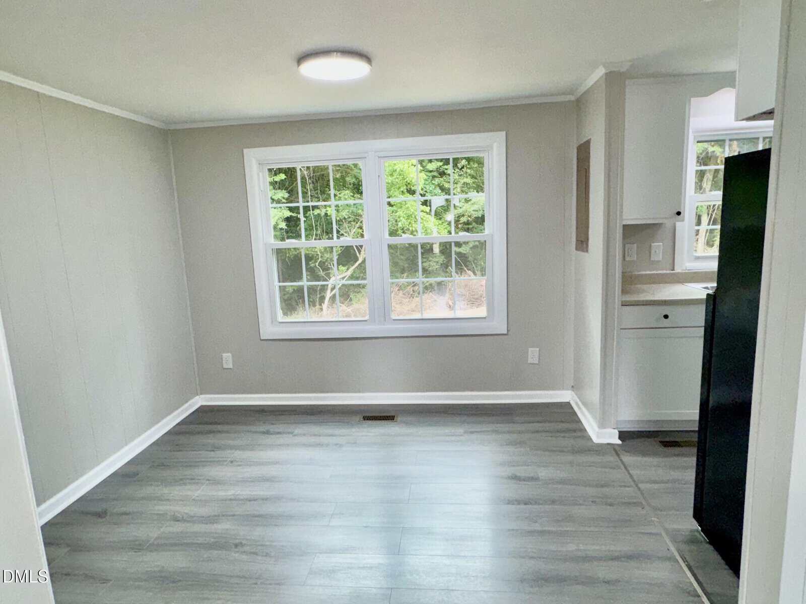 A small, empty room with light gray walls, wood-style flooring, and a large double window showing green trees outside. The corner of a kitchen with white cabinets and a black refrigerator is visible.