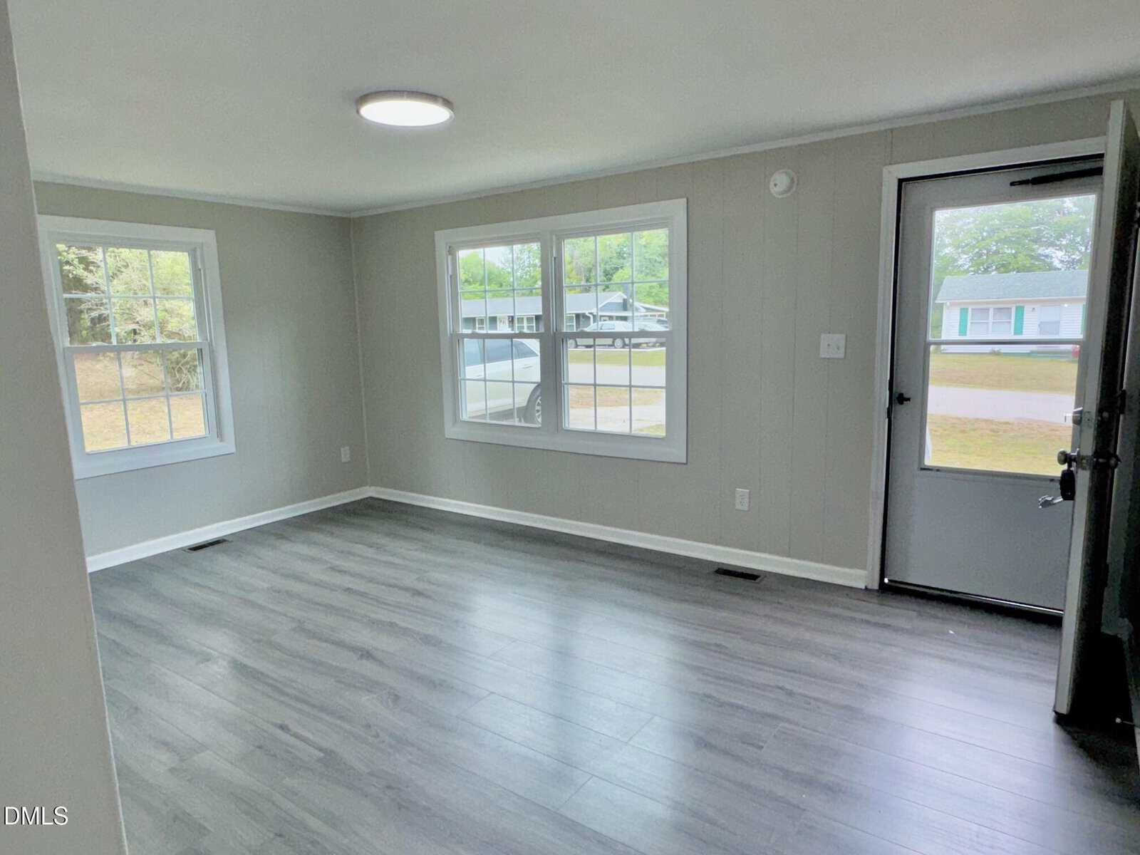 Bright, empty room with gray wood flooring, light gray walls, large windows letting in natural light, and a glass paneled door opening to a suburban street.