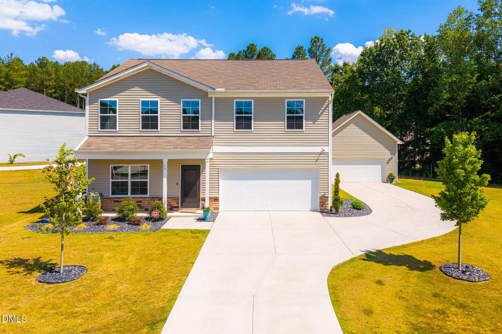 Two-story beige house with a three-car garage, neatly landscaped front yard, young trees, and a curved driveway, set against a backdrop of trees and a blue sky with scattered clouds.
