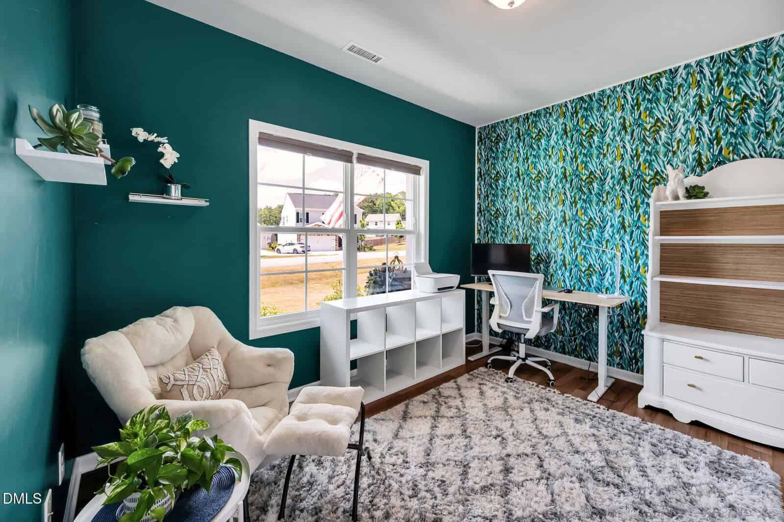A home office with teal and leaf-patterned walls, a large window, a white desk with a computer, a white chair, a cozy beige armchair, a leafy plant, and a white bookshelf on a patterned rug.