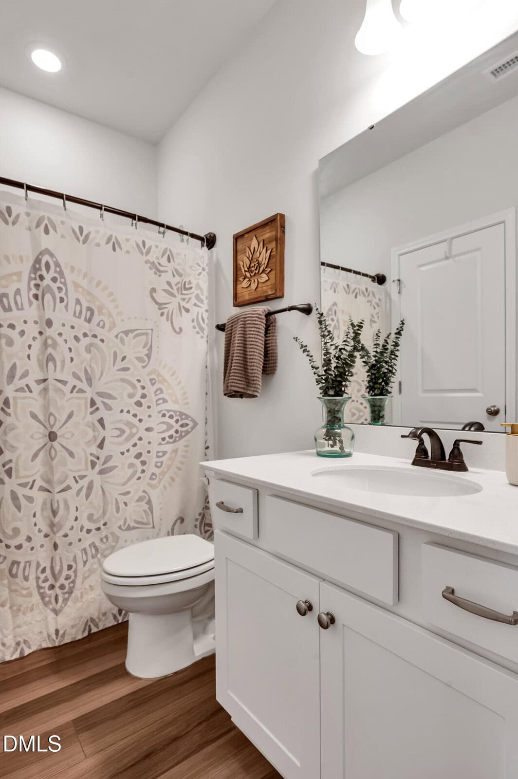 A bathroom with white cabinets, a white countertop, a toilet, a floral-patterned shower curtain, wood floors, decorative towels, a wooden wall art, and a vase with greenery on the sink.