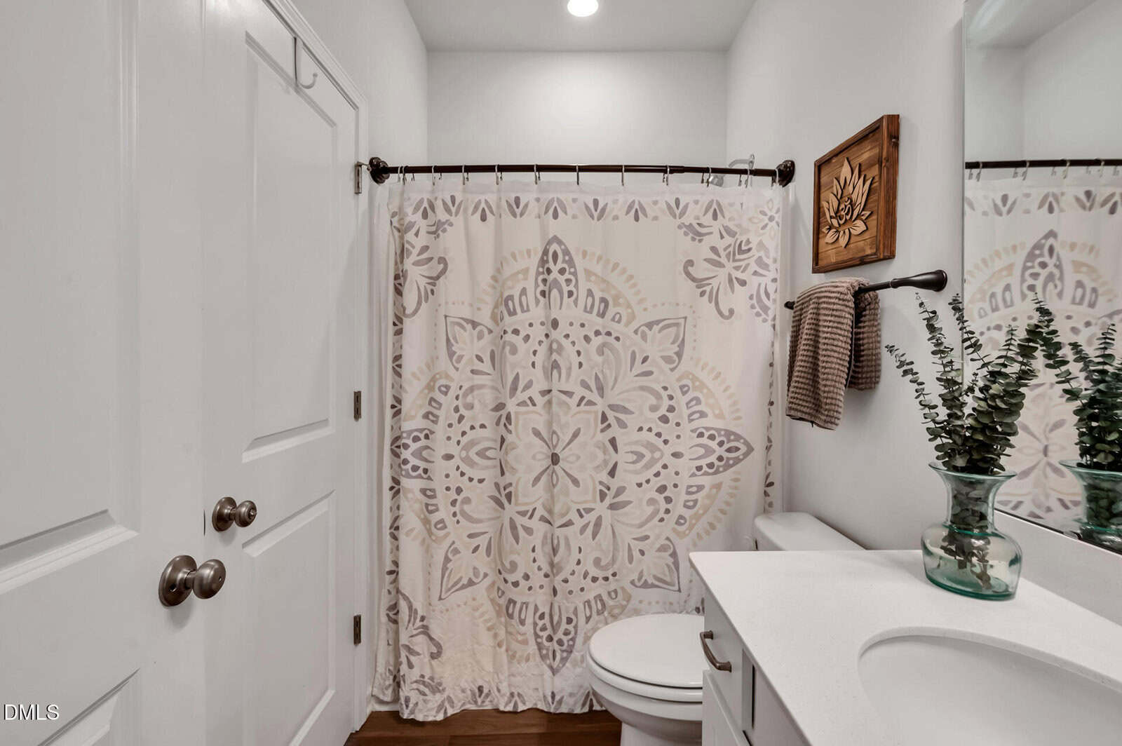 A small, white bathroom with a mandala-patterned shower curtain, white countertop, clear vase with greenery, towel rack with brown towels, and a framed wooden wall art.