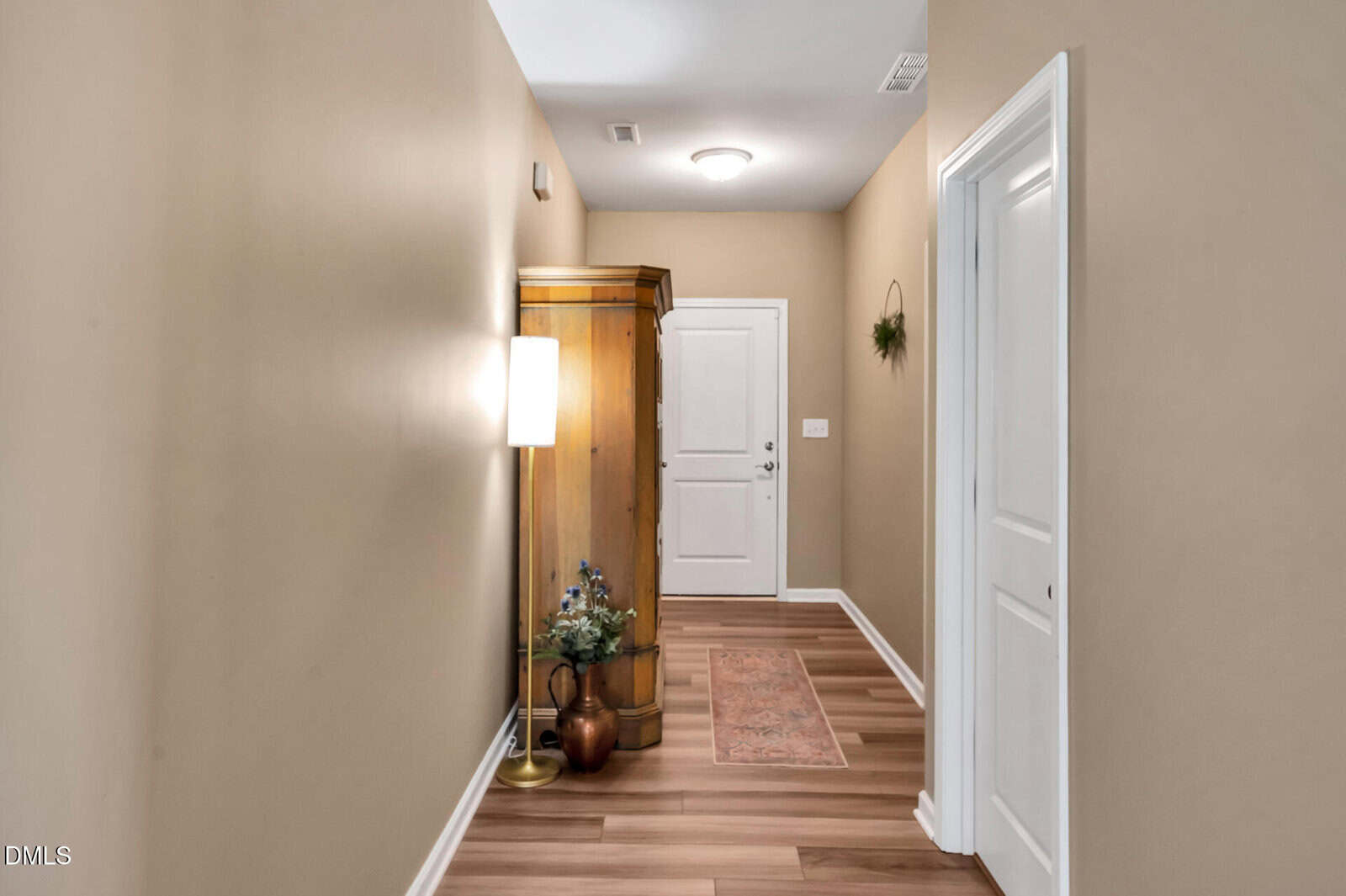A hallway with tan walls and wood flooring features a white door at the end, a wooden armoire, a floor lamp, a small potted plant, and a decorative rug on the floor.