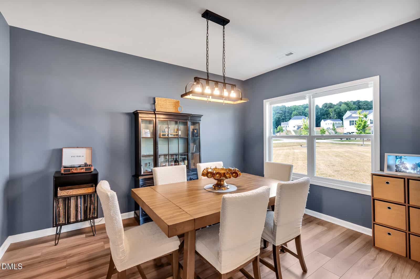 Modern dining room with a wooden table, six white chairs, a glass-front cabinet, a window showing a neighborhood, a record player, and wall shelves against blue-gray walls. A rectangular light fixture hangs above the table.