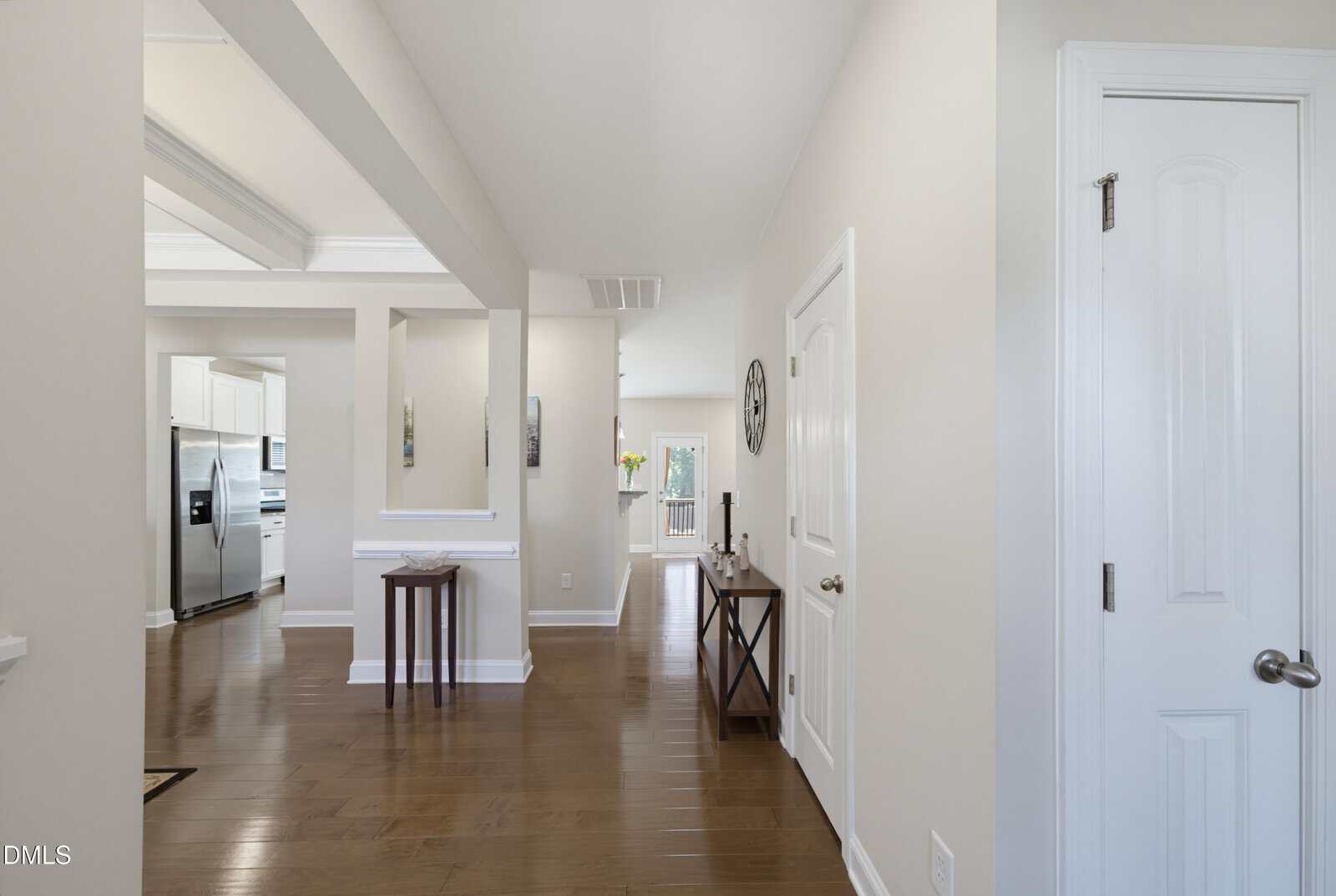 Bright hallway with hardwood floors, white walls, and doors. A small table with decor sits along the right wall, a console and mirror are farther back, and a modern kitchen is visible to the left.
