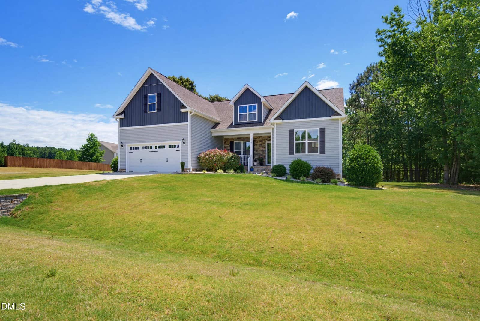 A modern two-story house with gray siding, white trim, and a double garage sits on a green lawn under a clear blue sky, with trees and shrubs surrounding the property.