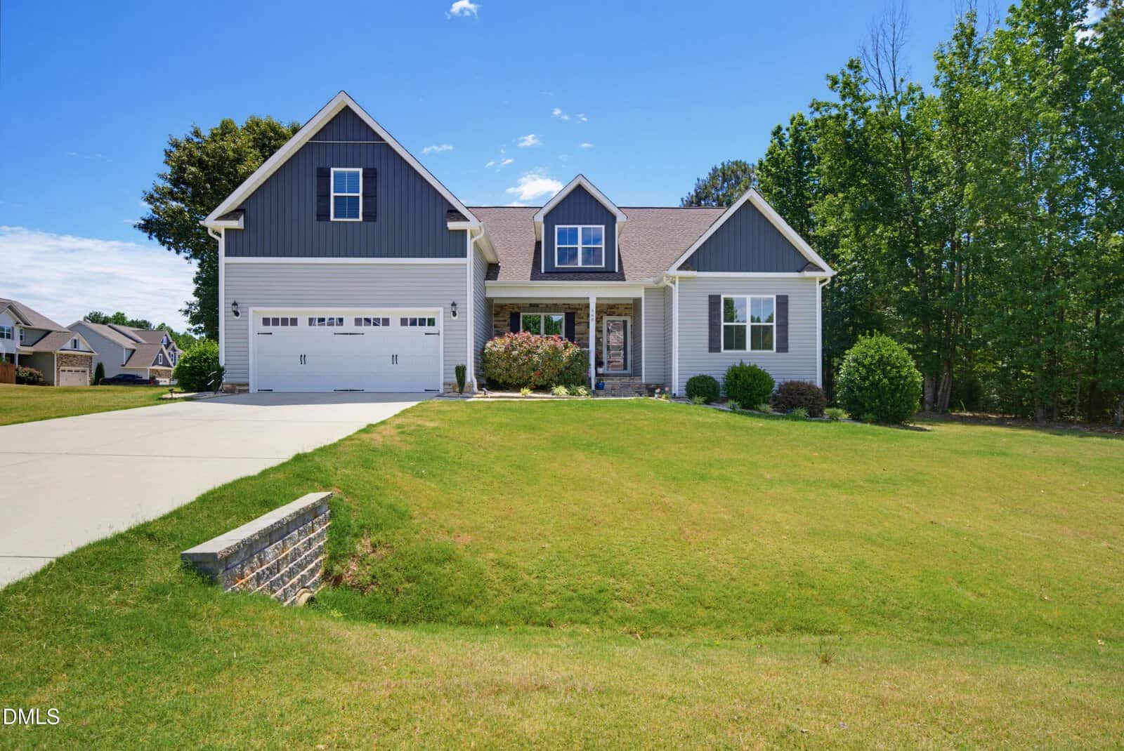 A modern two-story house with gray siding, white trim, and a two-car garage sits on a large, well-kept lawn with a few bushes and trees, under a clear blue sky.
