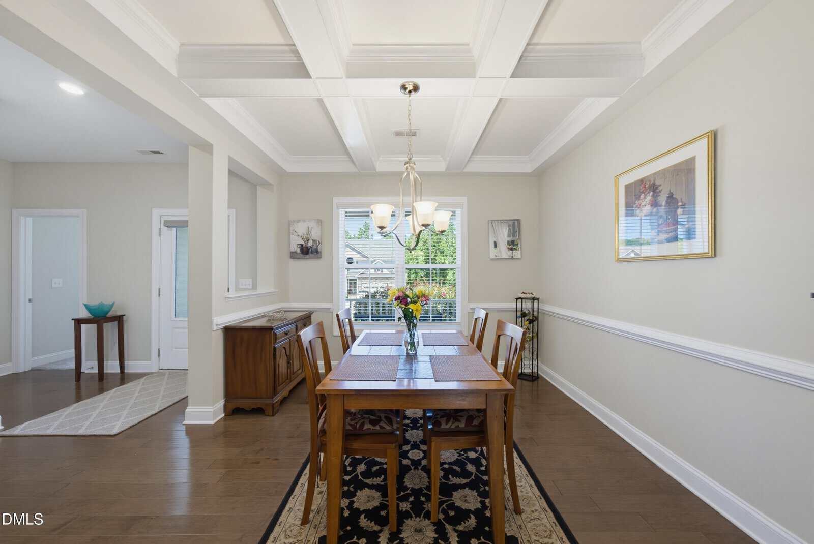 Bright dining room with a wooden table set for six, floral centerpiece, chandelier, artwork on beige walls, and large window letting in natural light. Hardwood floors and coffered white ceiling enhance the elegant space.