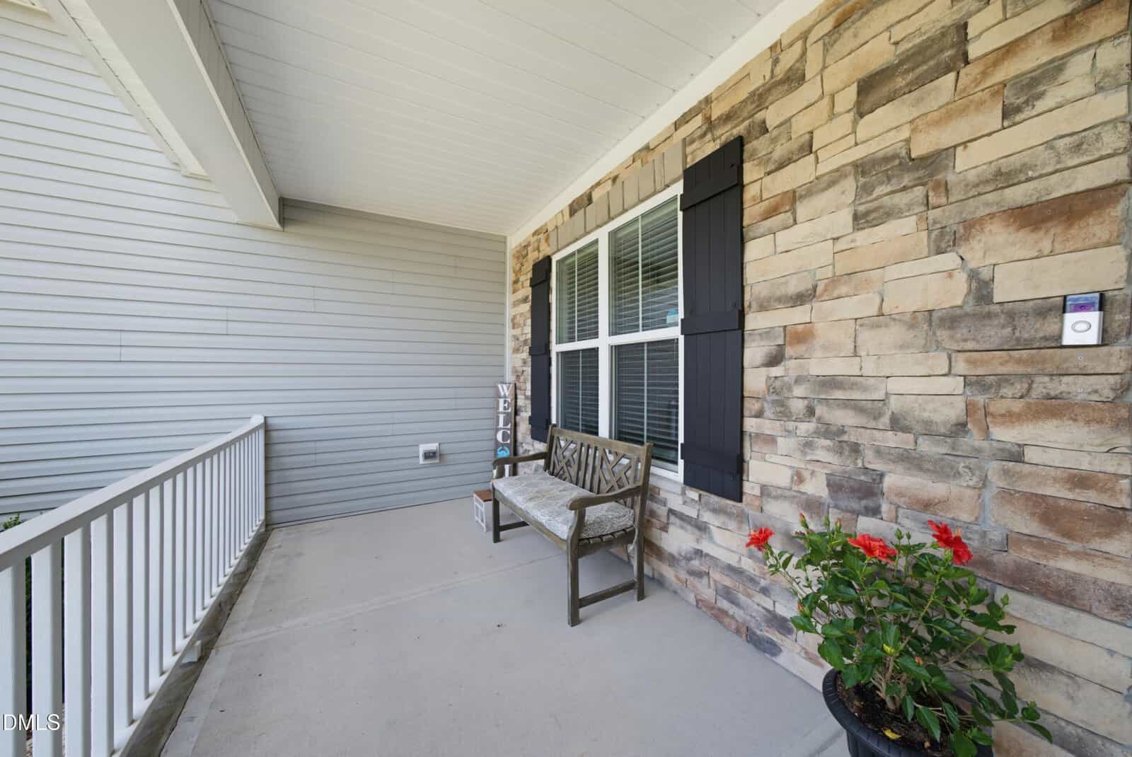 A front porch with a stone and siding exterior, a wooden bench with cushions, potted red flowers, and a Welcome sign by the window. White railing lines the porch.