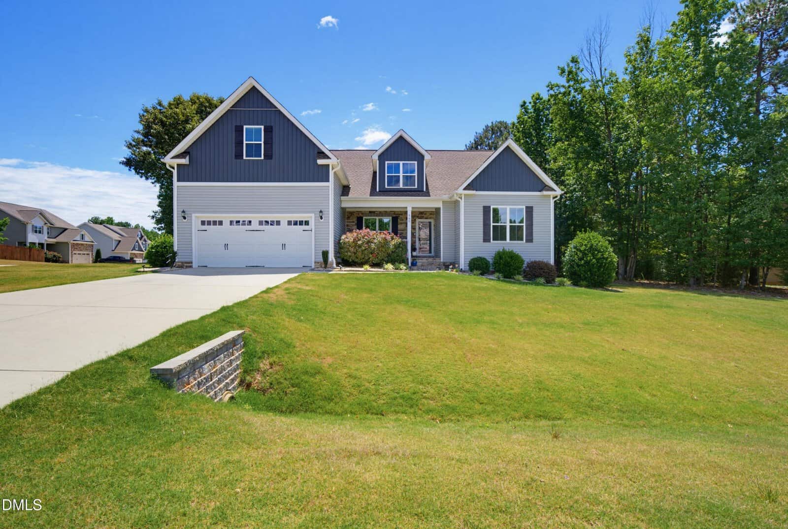 A modern suburban house with gray siding, white trim, a two-car garage, and a covered front porch, set on a large, well-manicured lawn bordered by trees under a bright blue sky.