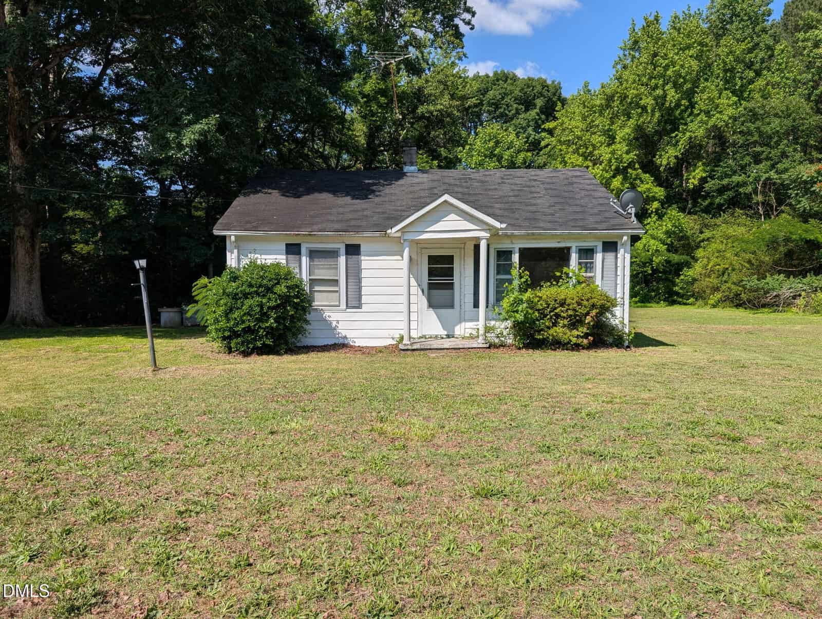A small white house with a dark roof, front porch, and green shrubs stands on a large grassy lawn at 1222 Serland Mill Road in Henderson. Tall trees and bushes surround the yard under a blue sky with scattered clouds.