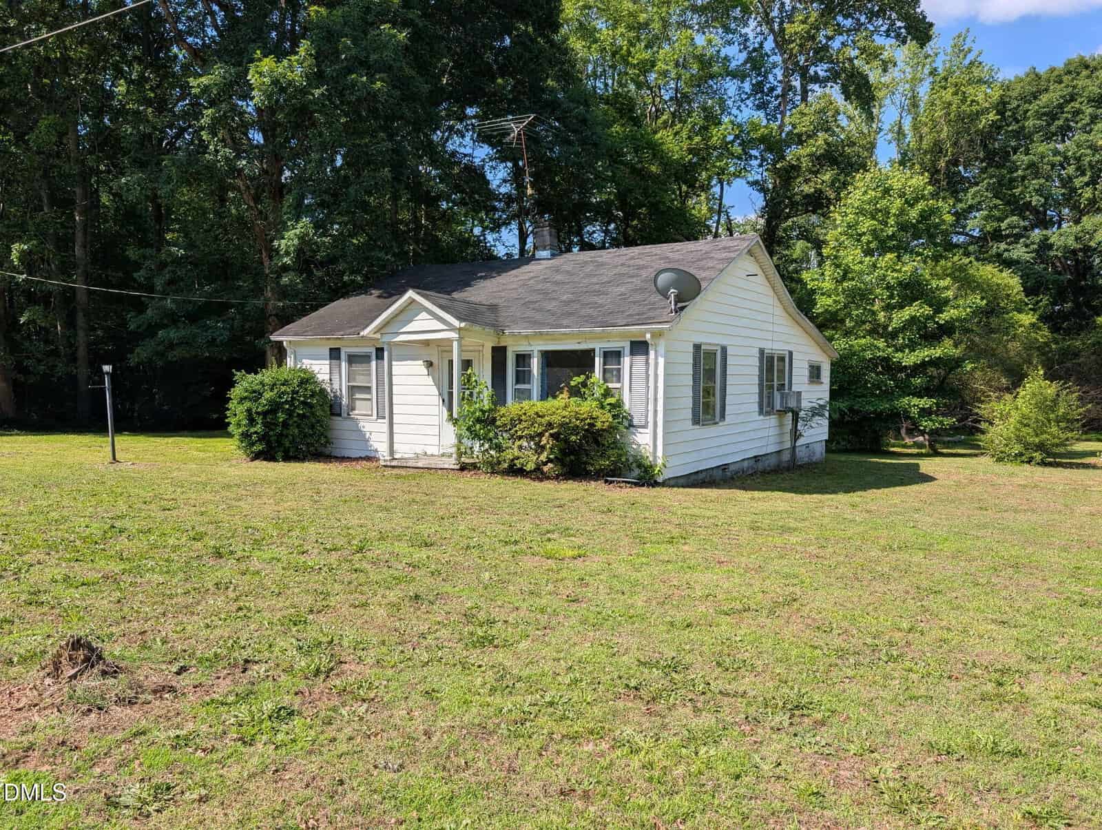 A small white single-story house with a dark roof, surrounded by grass and bushes, stands in a sunny yard with trees in the background. A satellite dish is attached to the side of the house.