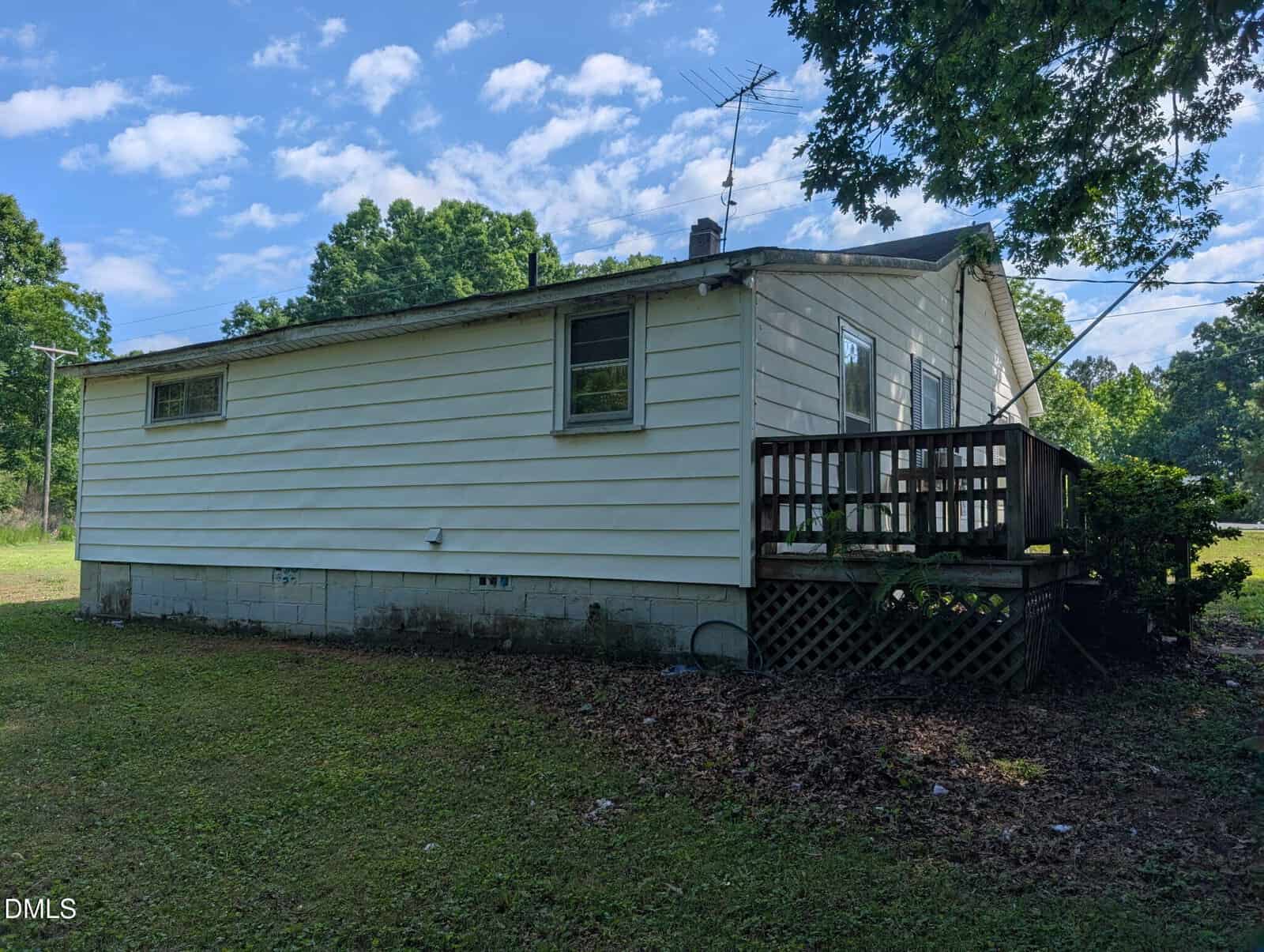 Small white house with horizontal siding, two windows, a wooden back deck, and steps leading down to a grassy yard. Trees and blue sky with scattered clouds are in the background.