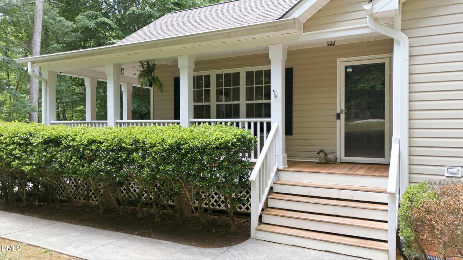 A beige house with a covered front porch, white railings, black shutters, and wooden steps leading up to the entrance. Bushes line the front, and trees are visible in the background.