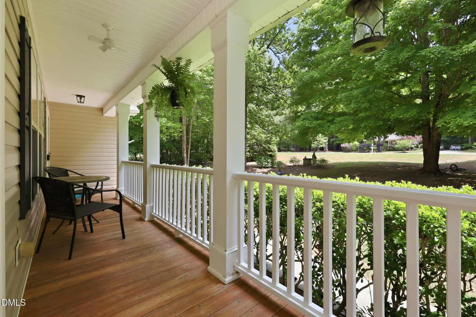 A wooden porch with white railings and columns features a small table and two chairs. Lush green trees and grass are visible in the yard beyond the porch. Hanging plants and lanterns add decorative touches.