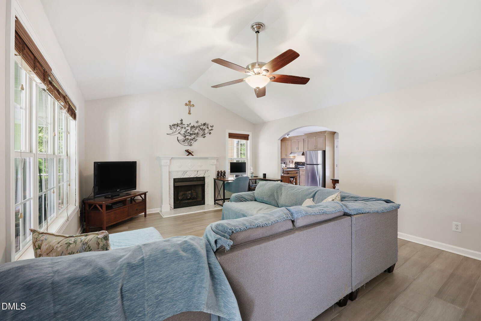 A bright living room with a sectional sofa, ceiling fan, TV on a wooden stand, and a fireplace with a mantel. Large windows line one wall, and the kitchen with stainless steel appliances is visible in the background.