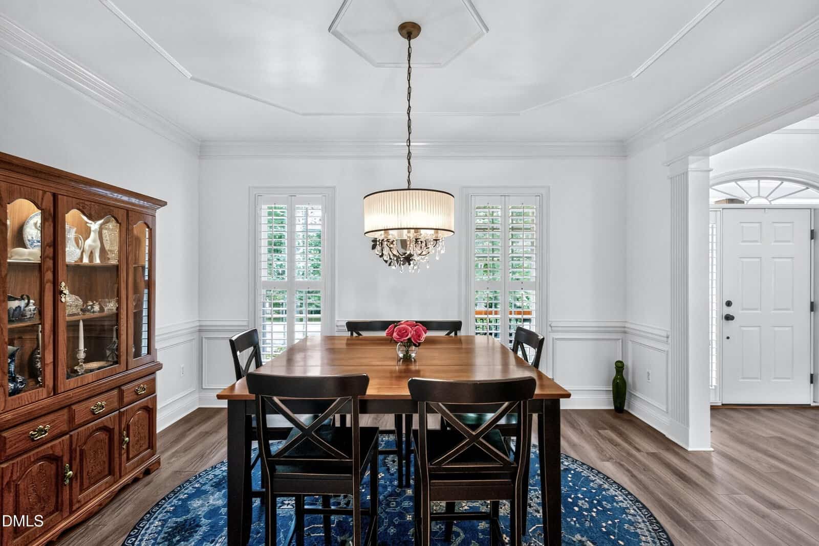 Bright dining room with a wooden table, six black chairs, a chandelier, blue round rug, and a china cabinet on the left. Two tall windows with shutters are behind the table; white walls and wood flooring complete the space.