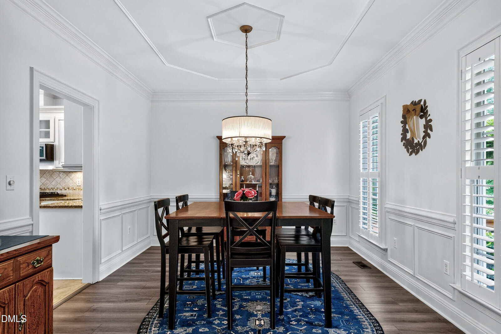 Elegant dining room with a dark wood table, six chairs, blue rug, chandelier, china cabinet, and white walls with wainscoting. Large windows with shutters allow natural light in.
