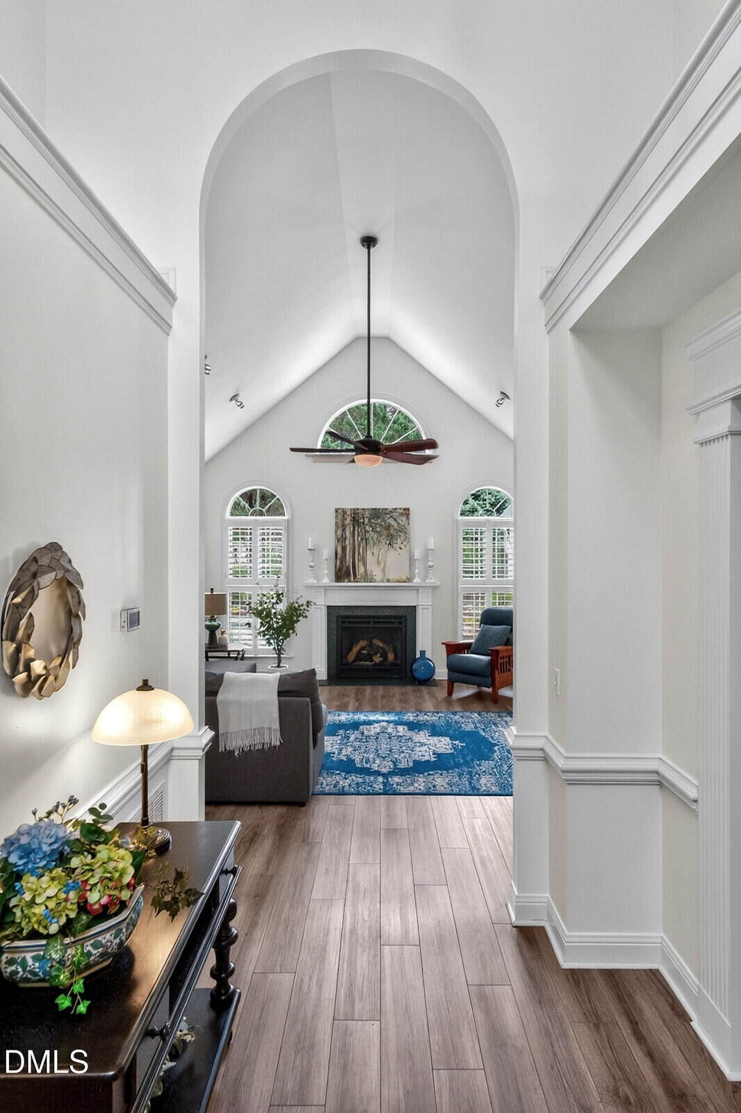 A hallway leads to a bright living room with vaulted ceilings, a fireplace, large windows, and cozy seating. A blue rug and decor accents add color, and natural light fills the space.