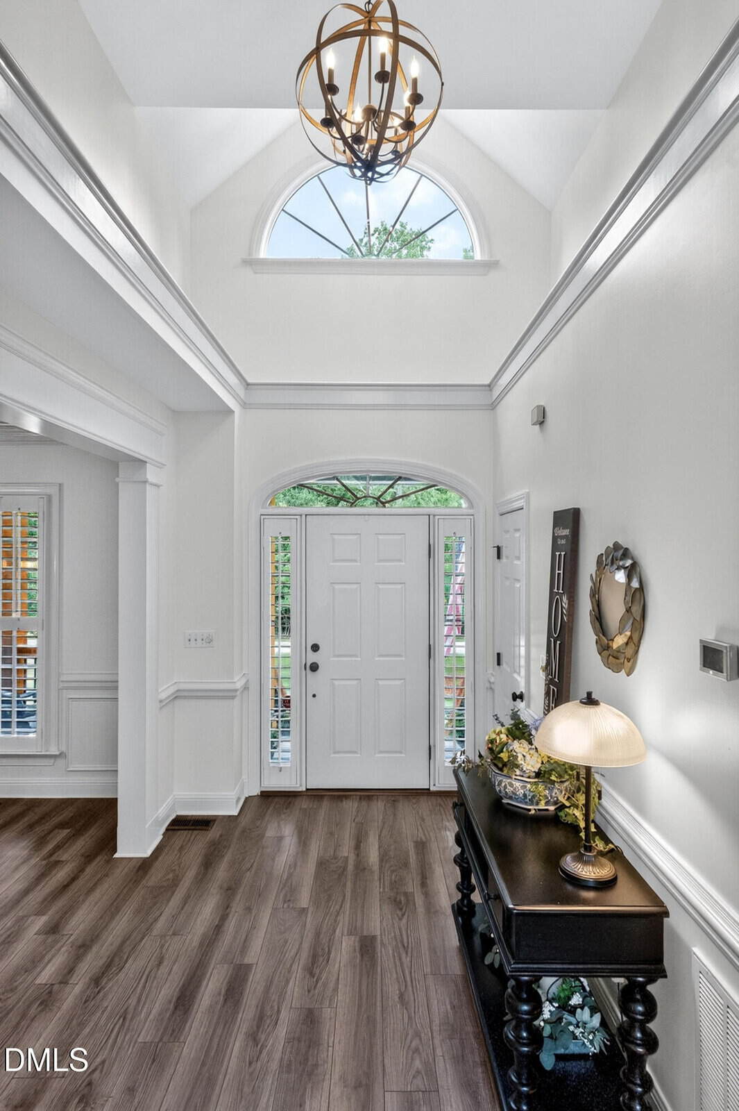 Bright entryway with tall ceilings, a large arched window above a white front door, wood floors, a black console table with decor and lamp, and a modern chandelier hanging overhead.