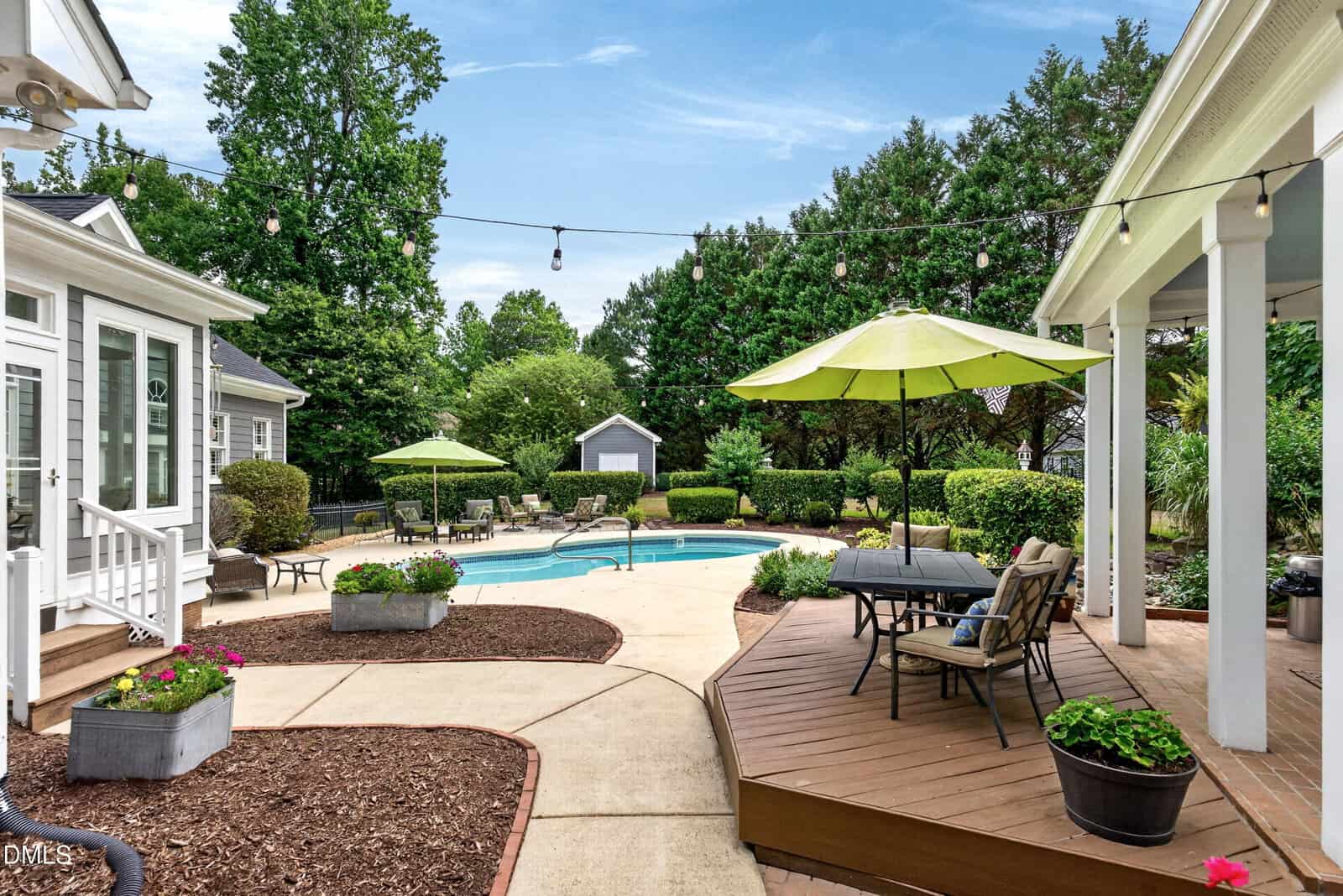 A backyard with a swimming pool, surrounded by a patio, deck with tables and chairs under green umbrellas, planters with flowers, string lights, and lush trees and bushes under a blue sky.