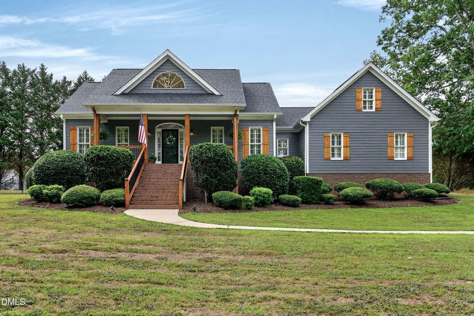 A gray house with wooden shutters and a covered front porch sits on a landscaped lawn with trimmed bushes, a curved walkway, and steps leading to the front door. Trees are in the background under a blue sky.