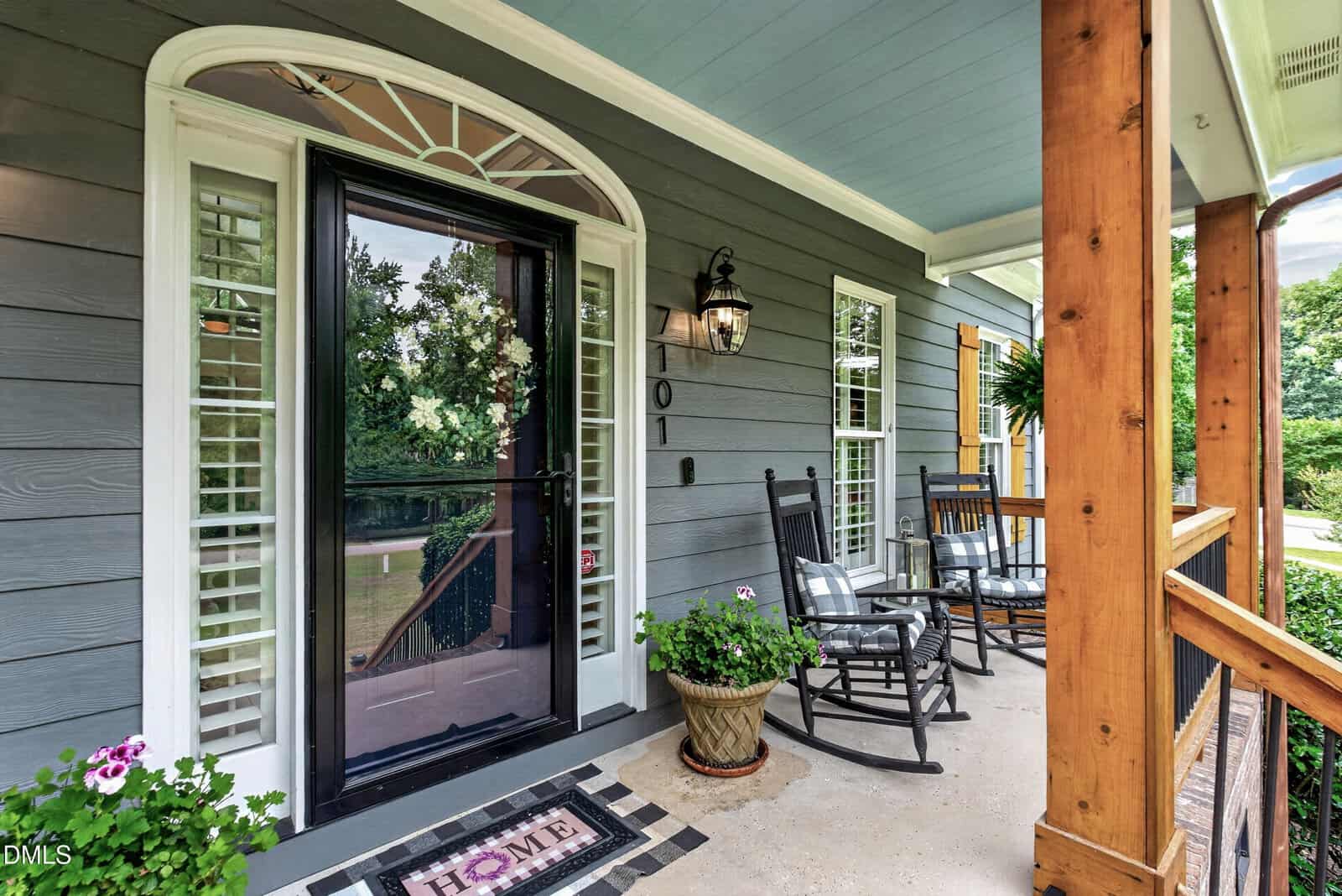 A cozy front porch with a glass door, two black rocking chairs, potted plants, a "HOME" doormat, wooden railings, and gray siding. Shutters frame the windows, and greenery is visible in the background.