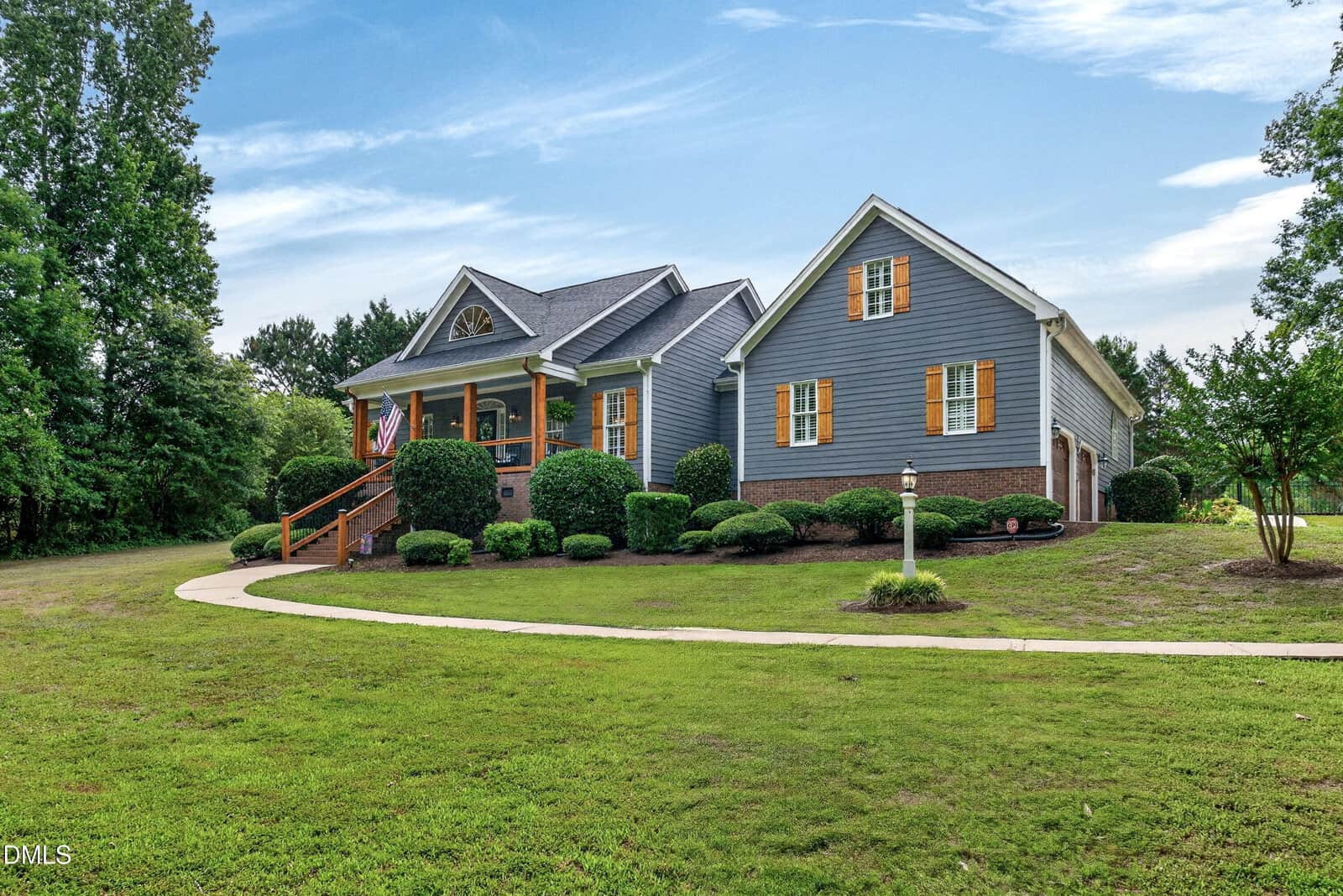A gray house with yellow shutters at 7101 Evelyn Circle in Zebulon features a front porch with an American flag, neatly trimmed bushes, a curved sidewalk, and a large green lawn surrounded by trees under a blue sky with light clouds.