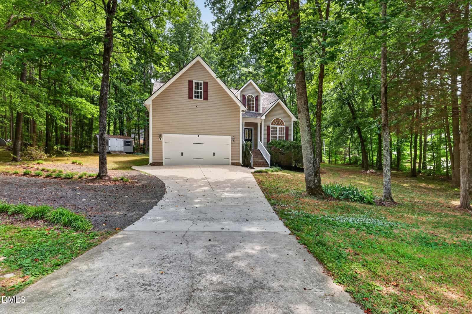 A beige two-story house with a double garage sits among tall trees at 4107 Maynard Circle in Franklinton. A concrete driveway leads to the home, and steps go up to the front entrance. The yard is green with grass and shaded by trees.