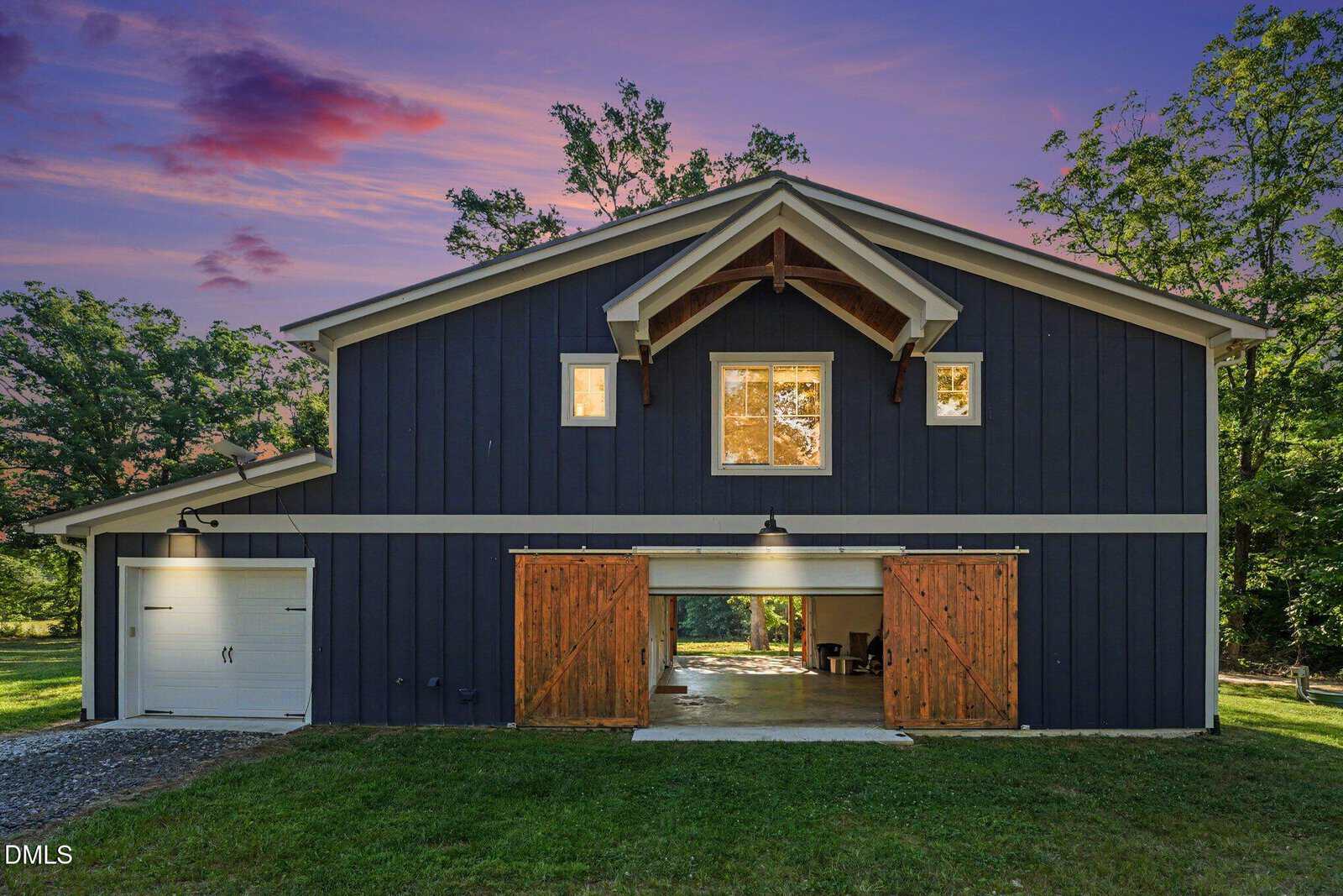 A modern dark blue barn-style building at sunset, with wooden double sliding doors open to reveal a lit interior, and a white garage door on the left side, surrounded by trees and a vibrant sky.