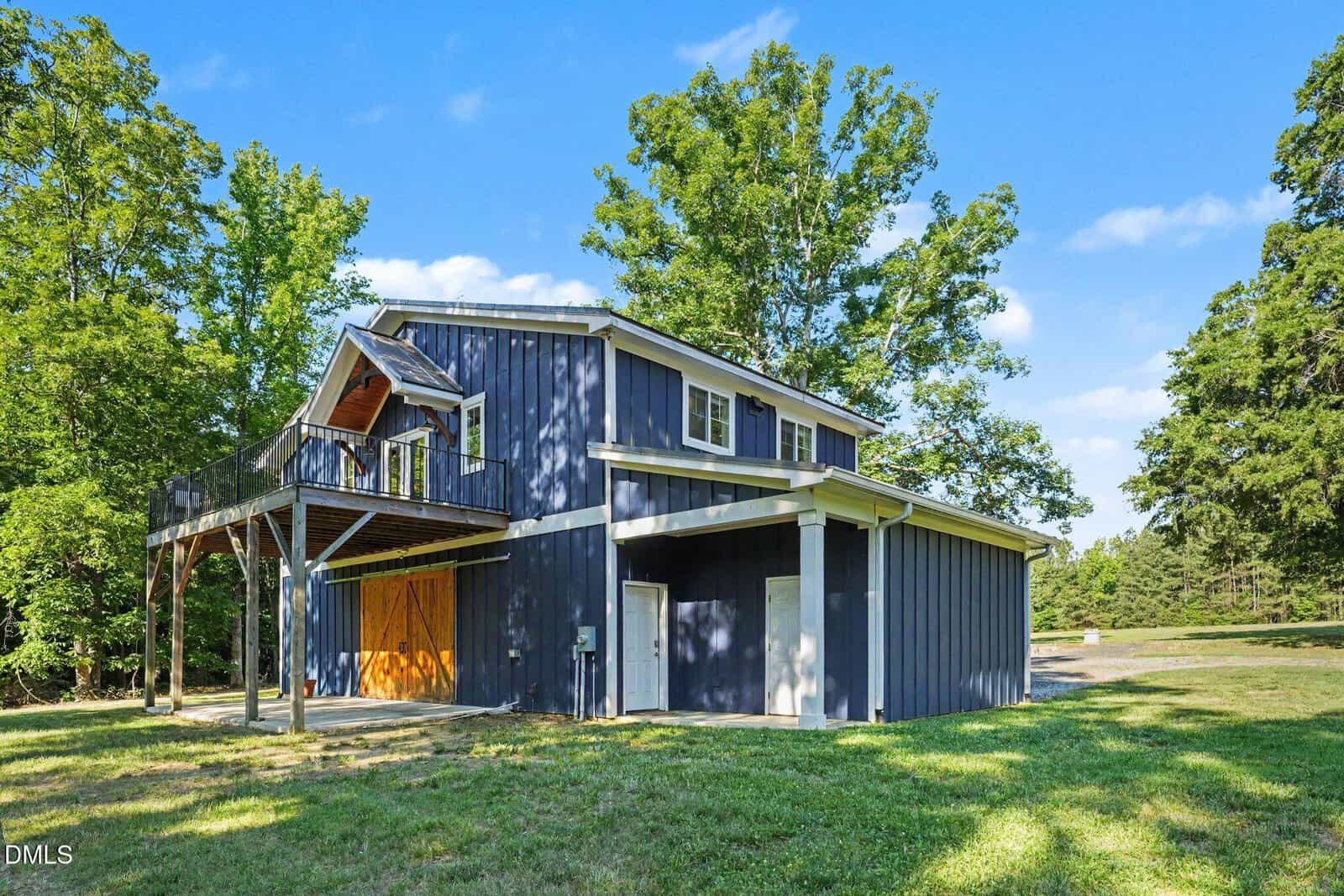 A modern two-story barn-style building painted dark blue with white trim, featuring a large balcony, wooden double doors, and surrounded by green grass and tall trees under a clear blue sky.