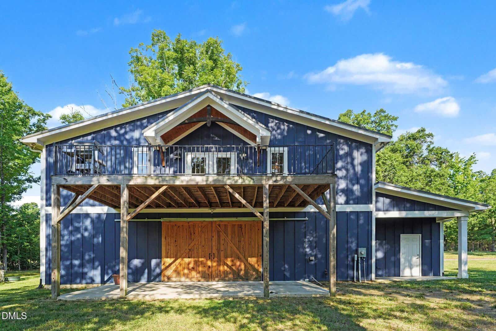 A large blue barn with white trim, wooden double doors, and an upper balcony with metal railings, surrounded by trees and a grassy area under a sunny sky.