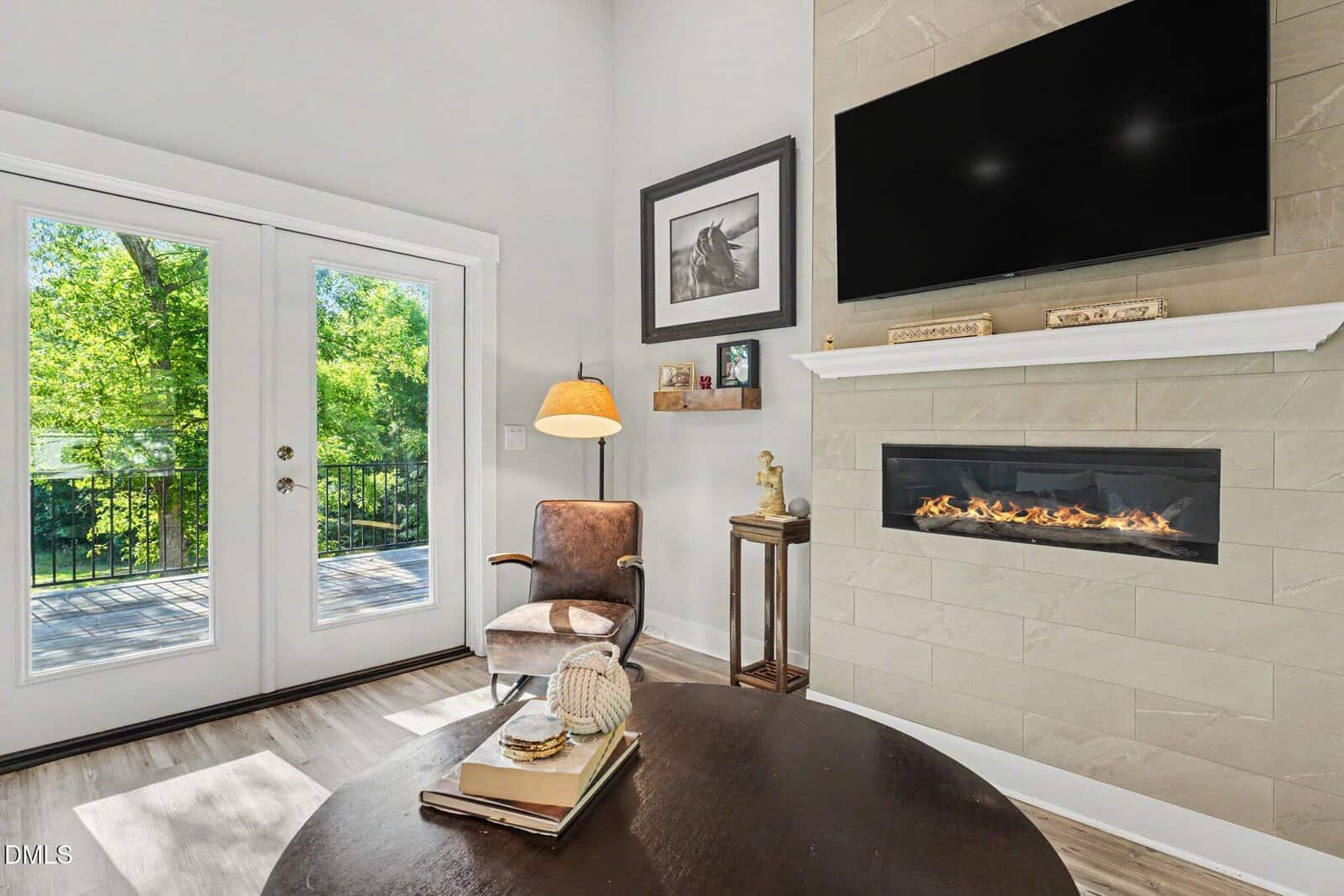 Bright living room with glass doors, a modern fireplace under a wall-mounted TV, an armchair, side table with decor, and a round coffee table. Trees and greenery are visible outside through the doors.