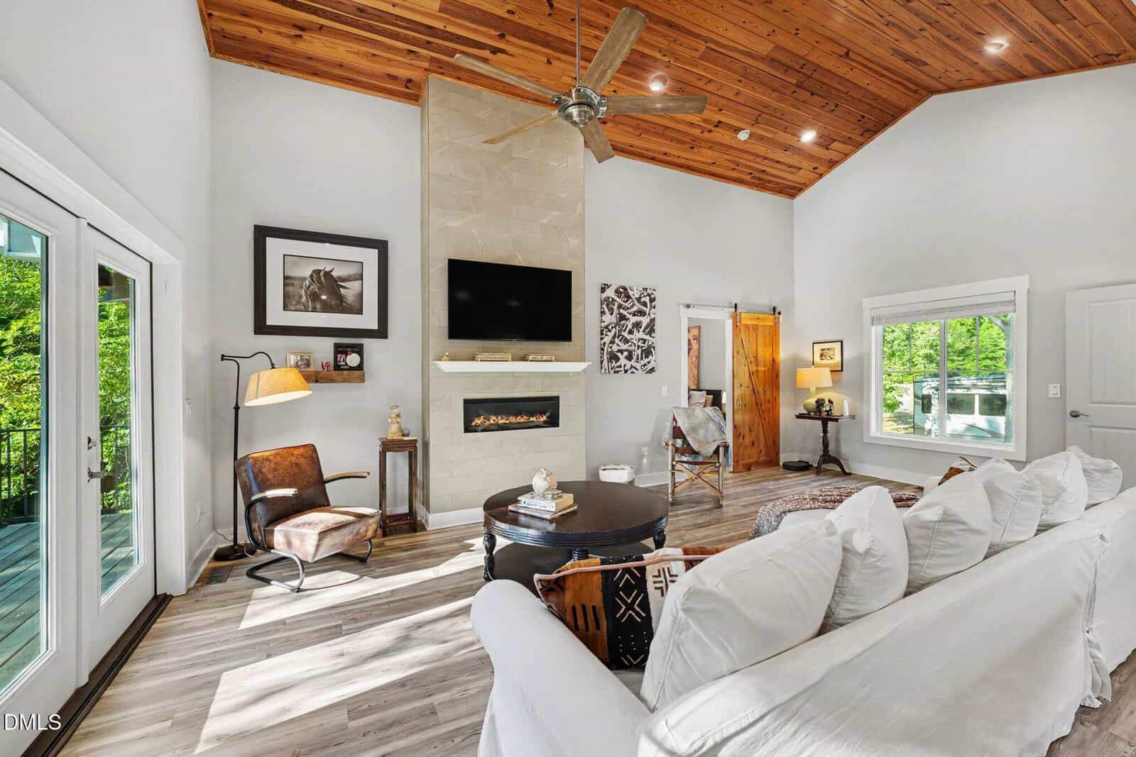 A bright living room with vaulted wood ceiling, white walls, a modern fireplace, wall-mounted TV, white sofa, round coffee table, and large windows overlooking greenery. Glass doors lead to a deck.