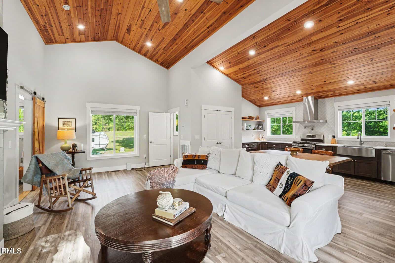 Bright, open living room and kitchen with wood-paneled ceilings, white walls, a white sofa, patterned pillows, round coffee table, and large windows overlooking greenery. Modern appliances and cozy decor complete the space.
