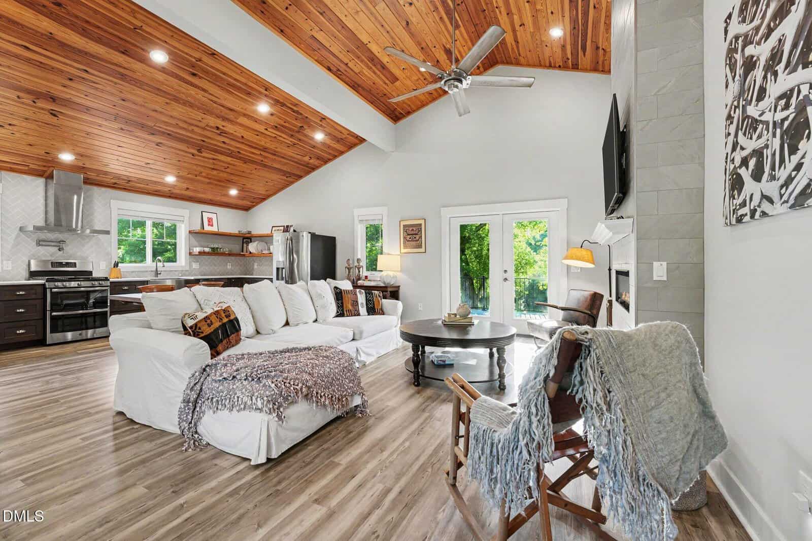 A cozy, modern living room with a white sofa, wooden ceiling, open kitchen, and large windows letting in natural light. A ceiling fan hangs above, and throws cover the sofa and chair. A round coffee table sits in the center.