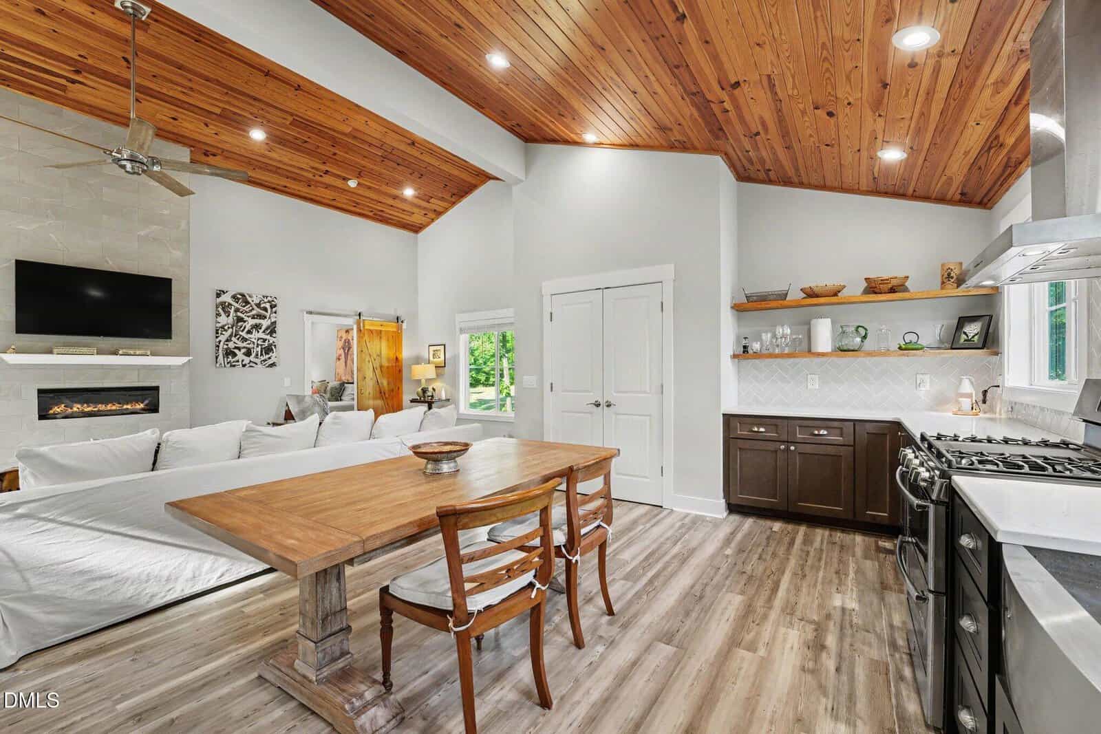Open kitchen and living space with wood plank ceiling, modern appliances, dark lower cabinets, open shelves, dining table with two chairs, white couch, wall-mounted TV, and bright natural light from windows.